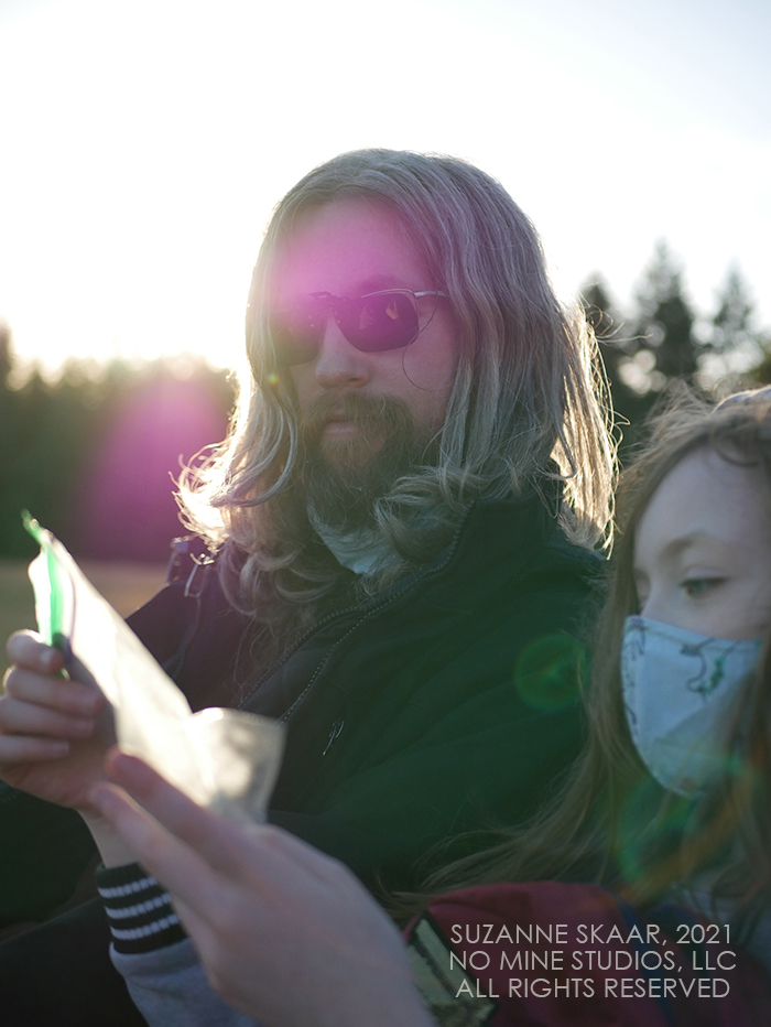 Father and child sitting next to each other in late evening. Sun glare behind father's head illuminates hair. Child is staring at snack in hand instead of looking at the camera.