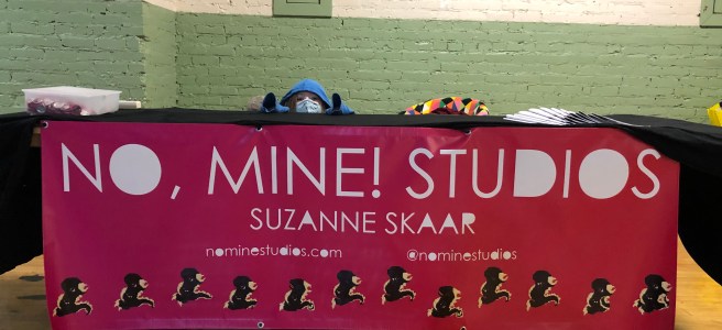 Picture of child in hoodie hiding behind table. On the front of the table is a large banner which reads "No, Mine! Studios; Suzanne Skaar; nominestudios.com; @nominestudios". A row of pixel art dinosaurs runs along bottom of banner.